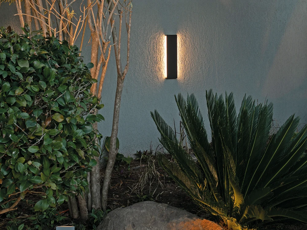 Arandela preta instalada junto ao jardim com luz quente sobre a parede